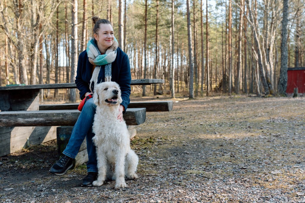 Else Verbeek sitting on a bench in a pine forest with a white dog beside her